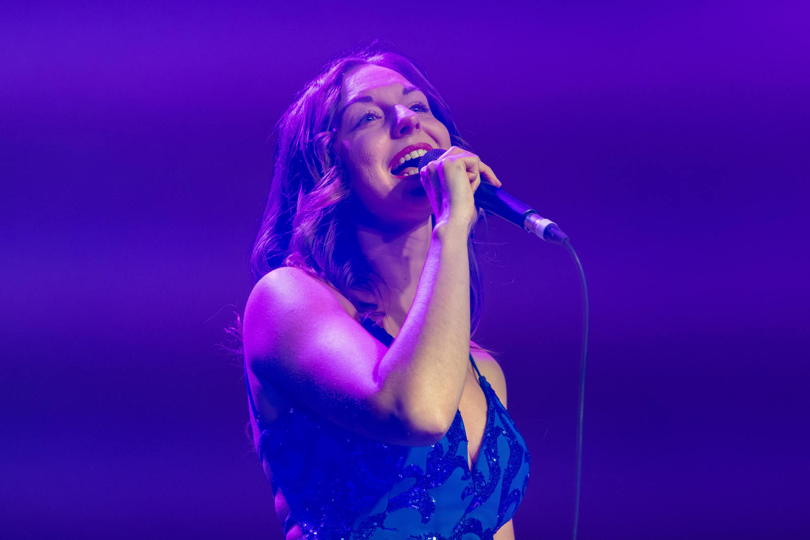 Sasha Kristoff singing into a handheld microphone in a sequined blue dress under purple stage light