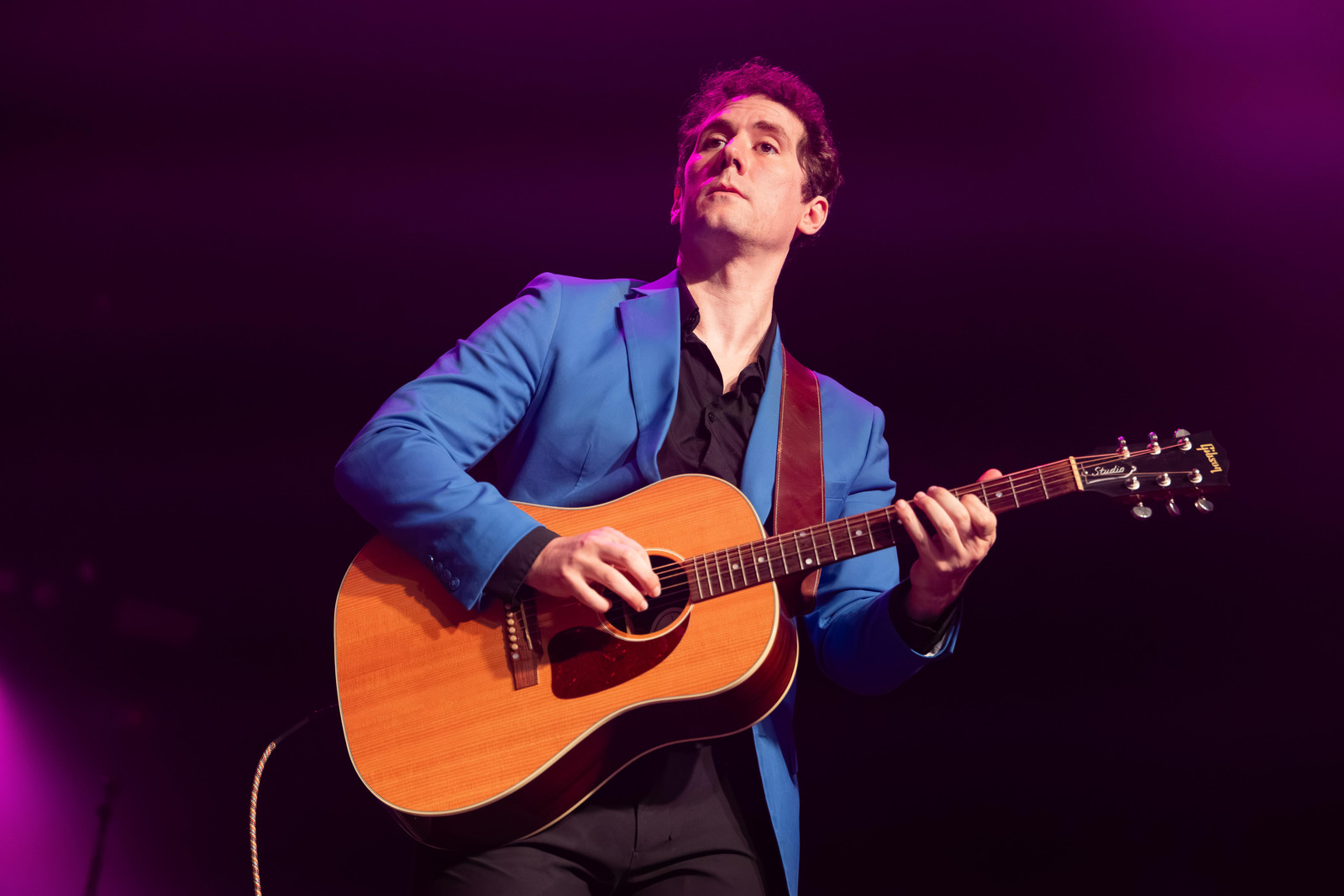 Stefan Schneider in a blue jacket playing an acoustic guitar under purple stage light